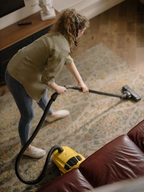 A person wearing full protective gear, including a white coverall, face mask, gloves, and shoe covers, is performing steam or deep cleaning on a beige carpet in a well-lit living room. The room featur